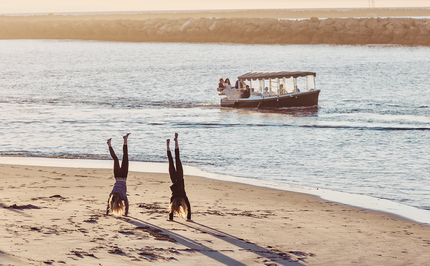 Two children doing handstands on the sand as an electric boat passes